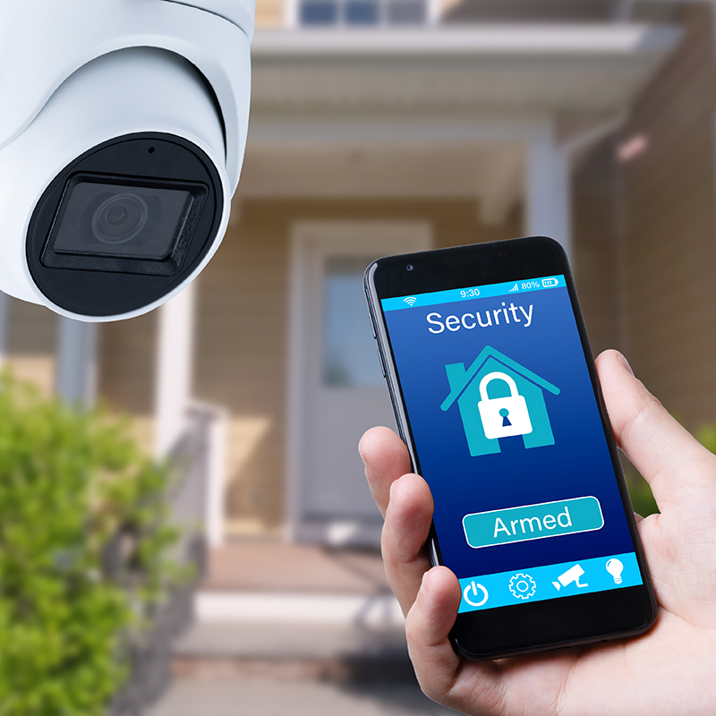 A security camera mounted outside a home paired with a smartphone displaying an armed security system app.